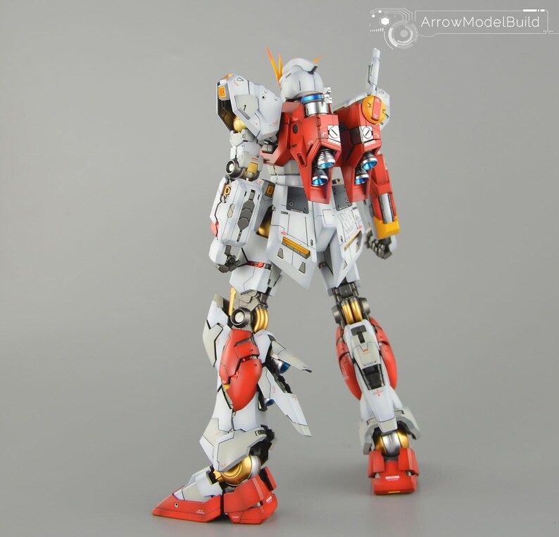 May include: A detailed model robot, predominantly white, red, and gold. The model features intricate mechanical detailing, including joints, panels, and weapons. The robot is posed with one leg forward, highlighting its design. The text "ArrowModelBuild" is visible.
