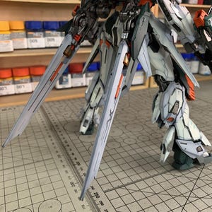 May include: A detailed, partially assembled model kit of a mech robot, featuring a complex design with gray, white, orange, and green accents. The model has large, stylized blades and intricate mechanical details, with the text "Arrow ModelBuild" visible.