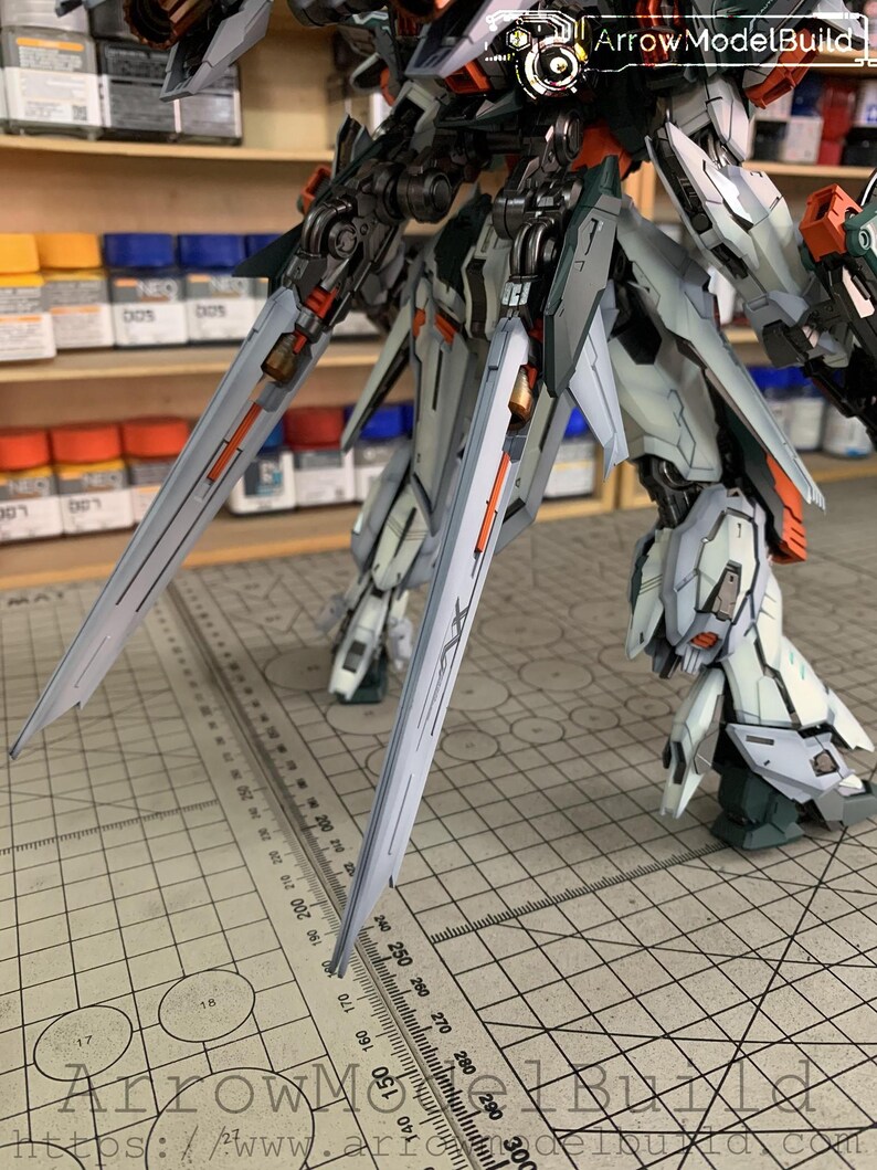 May include: A detailed, partially assembled model kit of a mech robot, featuring a complex design with gray, white, orange, and green accents. The model has large, stylized blades and intricate mechanical details, with the text "Arrow ModelBuild" visible.