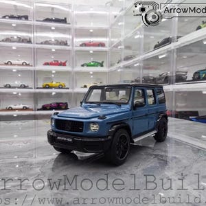 May include: A blue diecast model of a Mercedes-Benz G-Class SUV with black wheels and trim. The model is displayed in front of a collection of other model cars in clear display cases. The text "ArrowModelBuild" is visible.