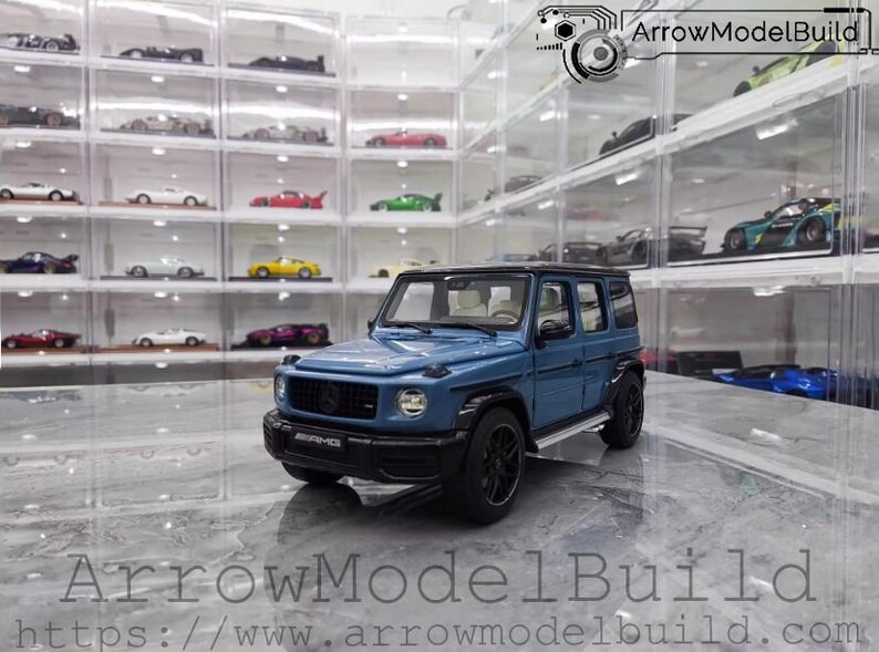 May include: A blue diecast model of a Mercedes-Benz G-Class SUV with black wheels and trim. The model is displayed in front of a collection of other model cars in clear display cases. The text "ArrowModelBuild" is visible.