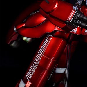May include: A detailed, red model kit of a mobile suit, featuring metallic accents and intricate mechanical details. The model has the text "STRUM BOOSTER SYSTEM" and "ModelBuild" on it. The kit is displayed against a black background.