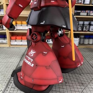 May include: A detailed, red and black model of a mobile suit robot, with visible panel lines and the text "CUSTOMIZE TYPE ZNABLE". The model features articulated legs and arms, and an orange pole. The model is set against a background of shelves with paint bottles.