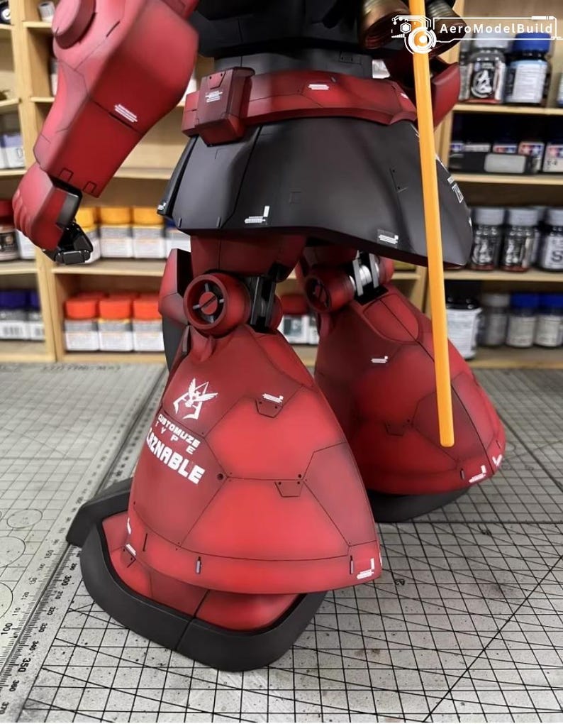 May include: A detailed, red and black model of a mobile suit robot, with visible panel lines and the text "CUSTOMIZE TYPE ZNABLE". The model features articulated legs and arms, and an orange pole. The model is set against a background of shelves with paint bottles.