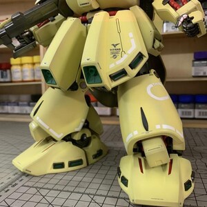 May include: A detailed, partially assembled model kit of a yellow and olive green mech. The model features intricate panel lines, green accents, and the "TITANS" logo. The image shows the lower body and legs of the model, with a gun visible.