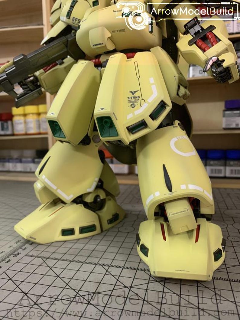 May include: A detailed, partially assembled model kit of a yellow and olive green mech. The model features intricate panel lines, green accents, and the "TITANS" logo. The image shows the lower body and legs of the model, with a gun visible.
