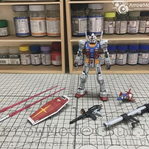以下が含まれることがあります： 赤、白、青、黄色の配色が特徴のガンダムロボットの精密模型キット。シールド、武器、ビームサーベルなどのアクセサリーが含まれています。模型は塗料の瓶とグリッド線のある表面を背景に配置されています。