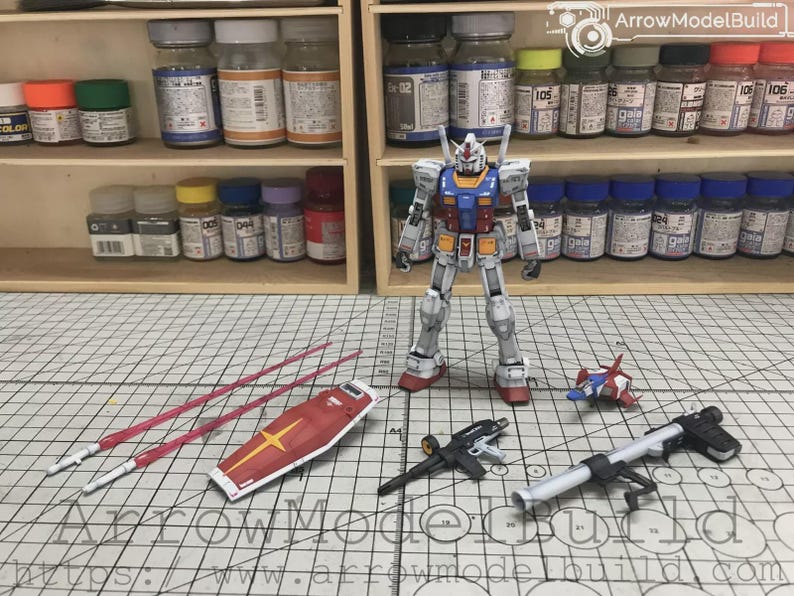 以下が含まれることがあります： 赤、白、青、黄色の配色が特徴のガンダムロボットの精密模型キット。シールド、武器、ビームサーベルなどのアクセサリーが含まれています。模型は塗料の瓶とグリッド線のある表面を背景に配置されています。