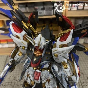 Puede incluir: Maqueta detallada de un robot mecha, con una combinaci&oacute;n de colores blanco, negro, rojo, dorado y azul. El robot tiene una armadura intrincada y estructuras en forma de alas. El texto "ArrowModelBuild" es visible en la esquina superior derecha.