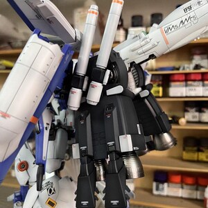 May include: A detailed model kit of a futuristic robot, featuring white, blue, and black armour with orange accents. The model has multiple thrusters and wings, with text that reads "EFSF" and "VMS/WrS". The AeroModelBuild logo is visible.