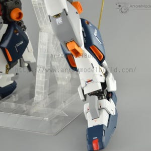May include: Close-up of a detailed model robot leg, featuring white, navy blue, gray, and orange accents. The model is mounted on a clear plastic stand. The text "ArrowModelBuild" and a website address are visible.