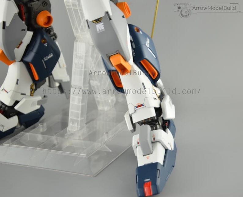 May include: Close-up of a detailed model robot leg, featuring white, navy blue, gray, and orange accents. The model is mounted on a clear plastic stand. The text "ArrowModelBuild" and a website address are visible.