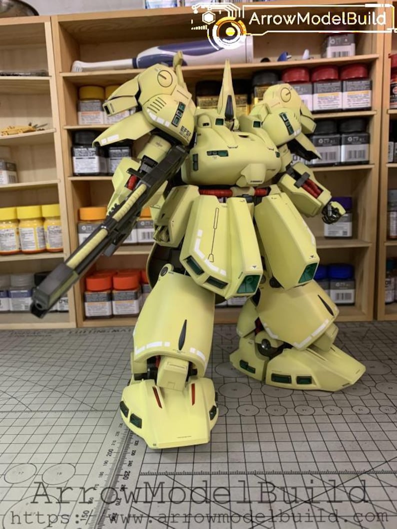 May include: A light yellow model robot with a large gun, red accents, and green details. The model is standing on a grid surface. The text "ArrowModelBuild" is visible in the image.