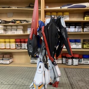 May include: A detailed model robot in red, black, white, and blue. The model has large wings and a long, red fin. It is positioned on a cutting mat, with paint bottles visible on shelves in the background.