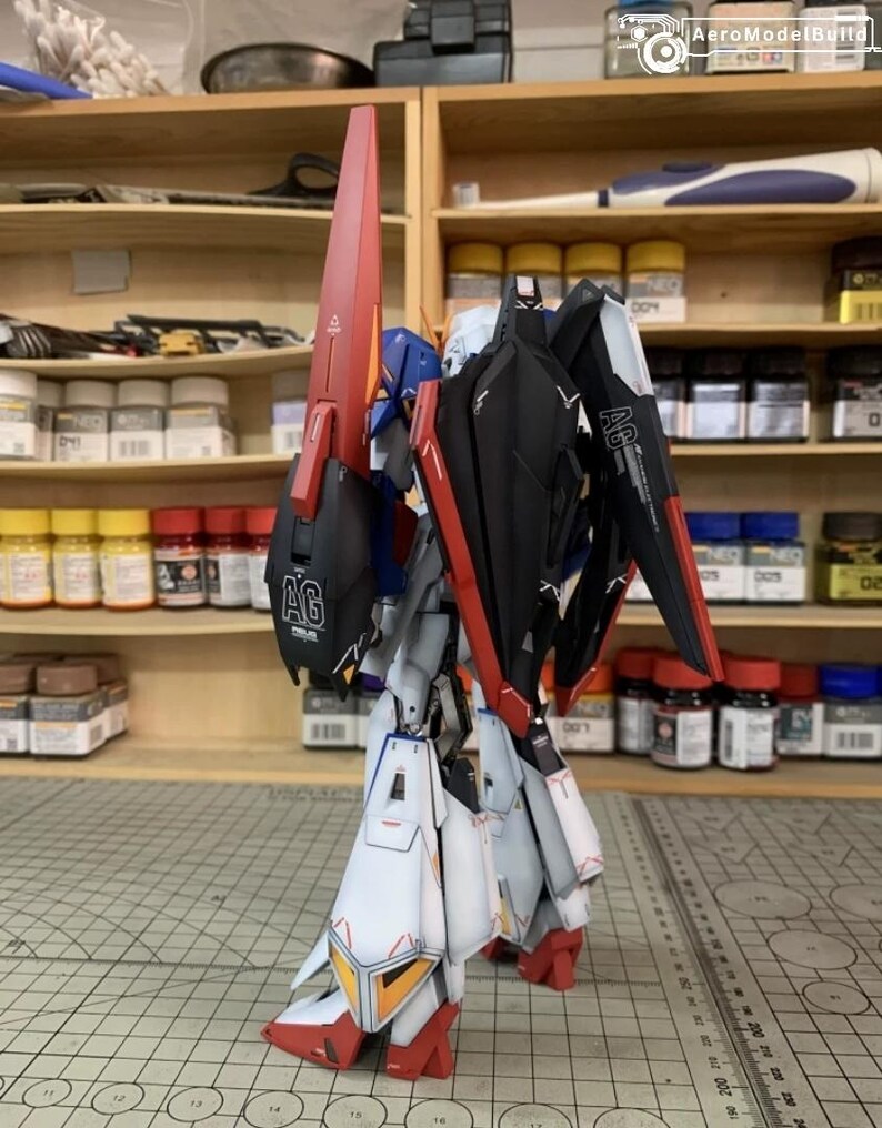 May include: A detailed model robot in red, black, white, and blue. The model has large wings and a long, red fin. It is positioned on a cutting mat, with paint bottles visible on shelves in the background.