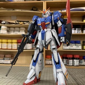 May include: A detailed model robot in blue, white, red, and black. The robot has a large rifle and a shield. It stands on a grid surface, with shelves and paint supplies in the background. The logo "AeroModelBuild" is visible.
