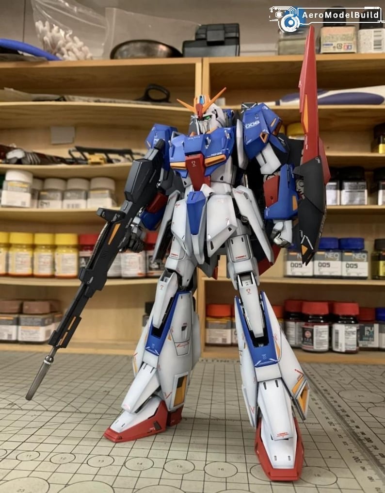 May include: A detailed model robot in blue, white, red, and black. The robot has a large rifle and a shield. It stands on a grid surface, with shelves and paint supplies in the background. The logo "AeroModelBuild" is visible.