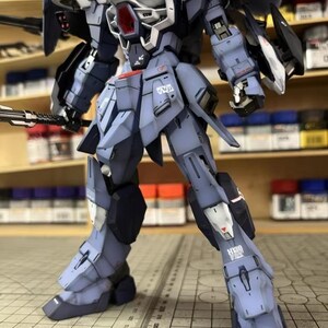 May include: A detailed model of a mecha robot, primarily in shades of blue and gray, with white and red accents. The robot is posed with a weapon in its left hand and has intricate mechanical details. The model is on a grid surface.