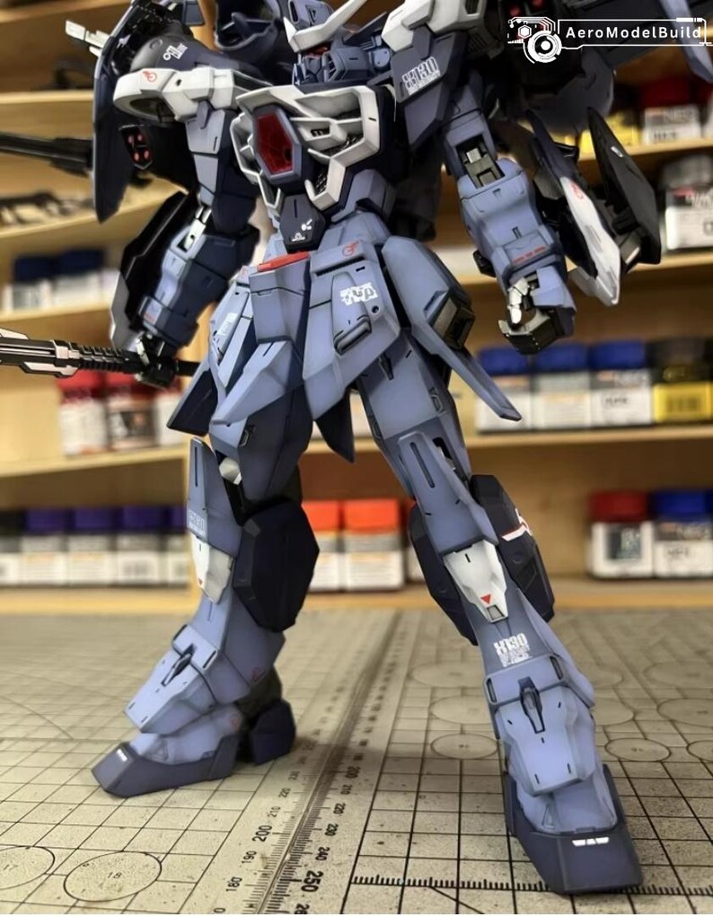 May include: A detailed model of a mecha robot, primarily in shades of blue and gray, with white and red accents. The robot is posed with a weapon in its left hand and has intricate mechanical details. The model is on a grid surface.