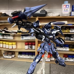 May include: A detailed, assembled model kit of a robot with a blue and gray color scheme. The robot is posed with its arms and legs spread, and it has a large weapon attached to its back. The background includes shelves with paint bottles and the text "AeroModelBuild".