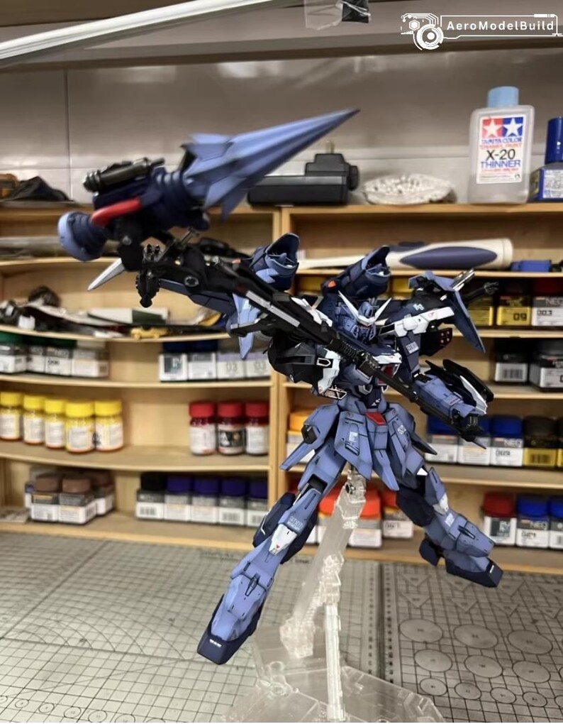 May include: A detailed, assembled model kit of a robot with a blue and gray color scheme. The robot is posed with its arms and legs spread, and it has a large weapon attached to its back. The background includes shelves with paint bottles and the text "AeroModelBuild".
