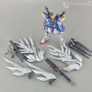 May include: A detailed model kit of a Gundam robot, featuring a blue, white, and red color scheme. The kit includes various components such as wings, weapons, and additional armor pieces. The model is posed on a gray surface.
