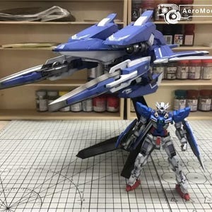 May include: A blue and white model of a futuristic aircraft with sharp, angular wings and a detailed robot figure. The robot is in a standing pose, with red and white accents. The model is on a grid-marked surface.