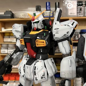May include: A detailed model of a Gundam robot, featuring a white, black, red, and yellow color scheme. The model has intricate details, including panel lines and markings. It is holding a black weapon and has a shield attached to its arm. The model is set against a blurred background.