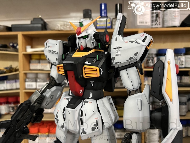 May include: A detailed model of a Gundam robot, featuring a white, black, red, and yellow color scheme. The model has intricate details, including panel lines and markings. It is holding a black weapon and has a shield attached to its arm. The model is set against a blurred background.