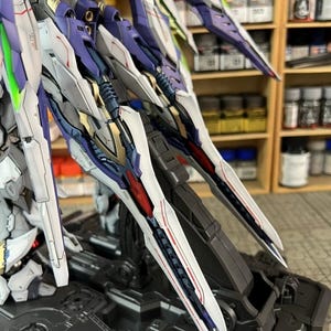 May include: A close-up of a model kit's mechanical wings, displaying intricate details. The wings are predominantly white, purple, red, and gold, with sharp, angular designs. The model is mounted on a grey base.