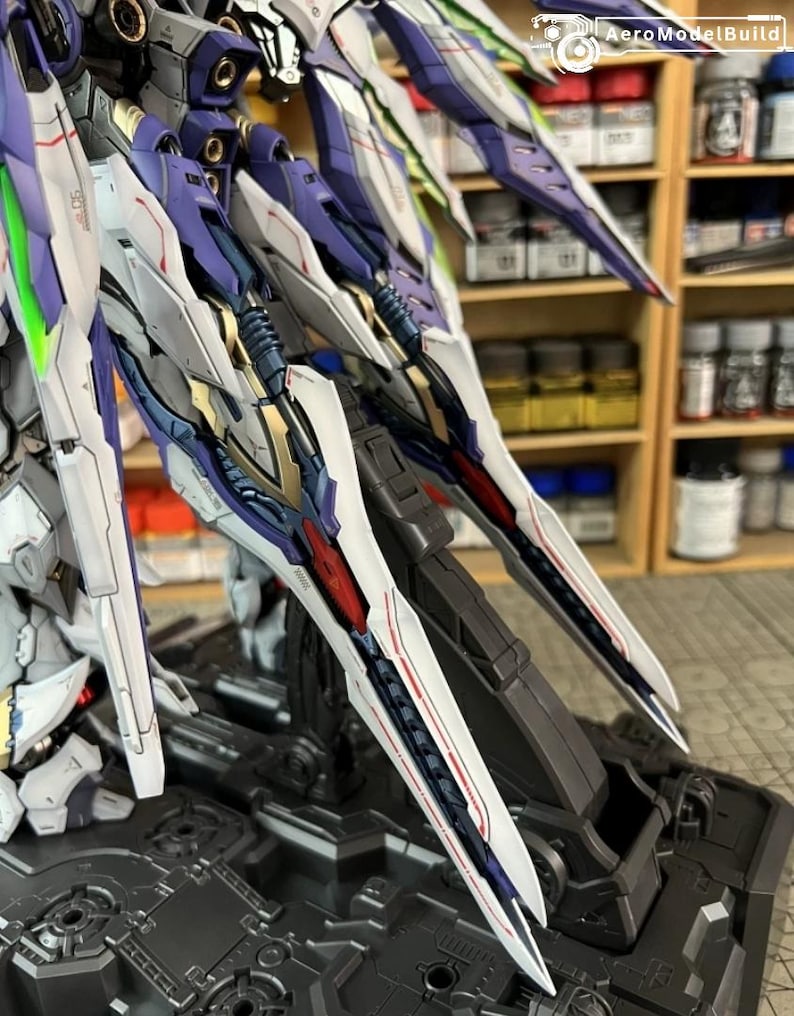 May include: A close-up of a model kit's mechanical wings, displaying intricate details. The wings are predominantly white, purple, red, and gold, with sharp, angular designs. The model is mounted on a grey base.