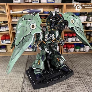 May include: A detailed model kit of a mech warrior with large, articulated wings. The figure is primarily olive green with black and gold accents. It stands on a black base, and the background shows shelves with paint bottles.