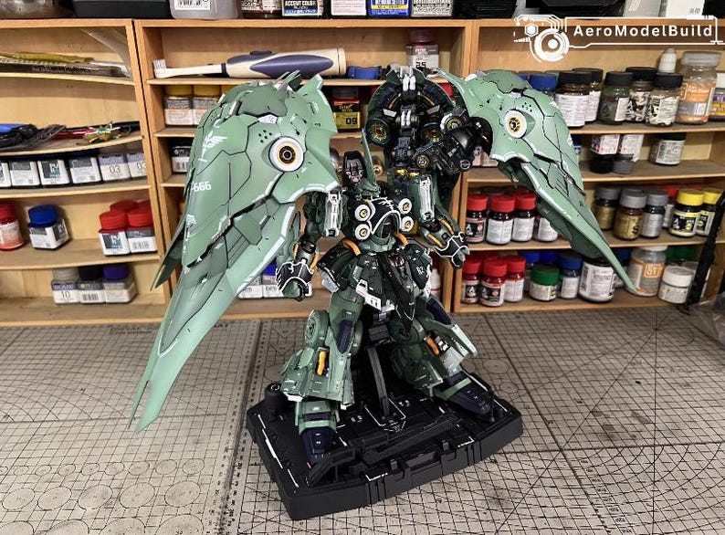 May include: A detailed model kit of a mech warrior with large, articulated wings. The figure is primarily olive green with black and gold accents. It stands on a black base, and the background shows shelves with paint bottles.