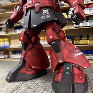 May include: A detailed, red and black model of a mobile suit robot. The model features intricate panel lines, decals, and the text "PRINCIPALITY OF ZEON". The robot's legs and lower body are in focus, showcasing the mechanical design.