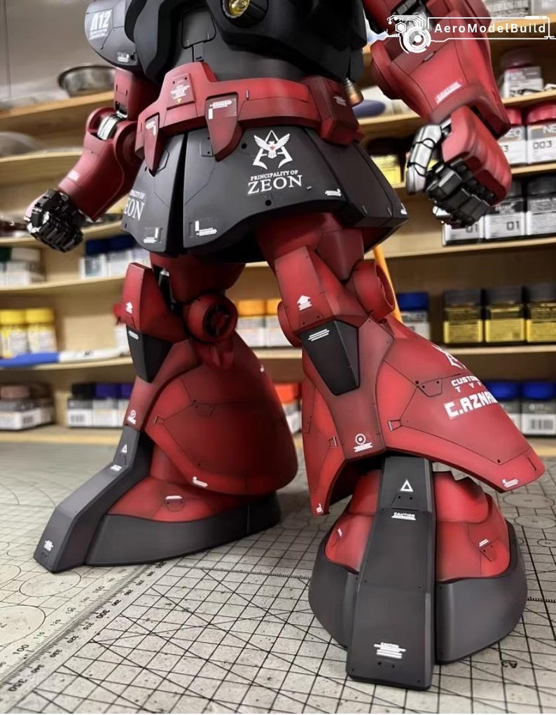 May include: A detailed, red and black model of a mobile suit robot. The model features intricate panel lines, decals, and the text "PRINCIPALITY OF ZEON". The robot's legs and lower body are in focus, showcasing the mechanical design.