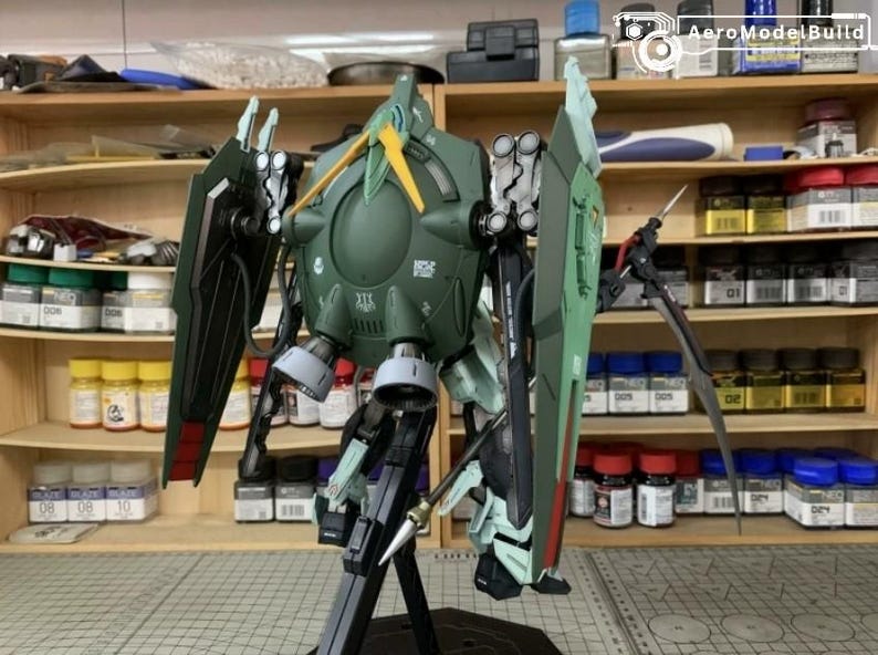 Puede incluir: Modelo detallado de un robot mec&aacute;nico verde y negro con grandes escudos y armas. El modelo est&aacute; sobre un soporte negro, con un dise&ntilde;o detallado. El fondo muestra estantes con botes de pintura y herramientas.