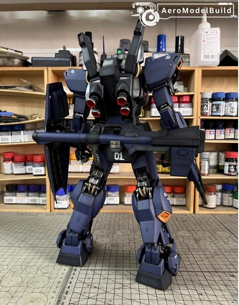 Pu&ograve; includere: Modello dettagliato di un robot mecha blu scuro e nero, con un grande fucile e pi&ugrave; propulsori. Il modello ha accenti arancioni e il numero "01" &egrave; visibile. Il logo AeroModelBuild &egrave; nell'angolo in alto a destra.