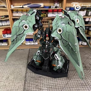 May include: A detailed model kit of a mech robot, predominantly green and black, with large, articulated wings. The model is on a black base, showcasing intricate panel lines and mechanical details. The background includes shelves with model-building supplies.