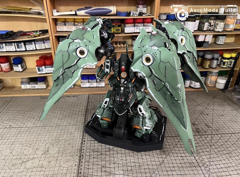 May include: A detailed model kit of a mech robot, predominantly green and black, with large, articulated wings. The model is on a black base, showcasing intricate panel lines and mechanical details. The background includes shelves with model-building supplies.