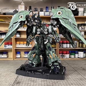 May include: A detailed model of a mecha robot with large, articulated wings. The robot is primarily dark green with black and orange accents. The model is on a black base, with the text "AeroModelBuild" in the top right corner.