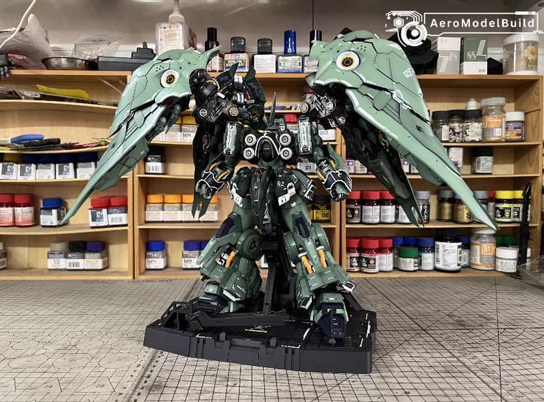 May include: A detailed model of a mecha robot with large, articulated wings. The robot is primarily dark green with black and orange accents. The model is on a black base, with the text "AeroModelBuild" in the top right corner.