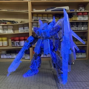 May include: A transparent blue model robot with large weapons and armor. The robot is standing on a gray surface with a grid pattern. The background includes shelves with various bottles and containers.
