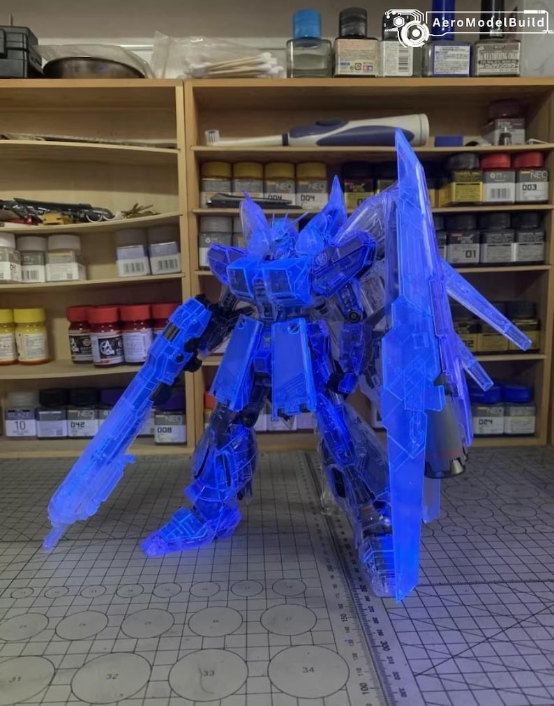 May include: A transparent blue model robot with large weapons and armor. The robot is standing on a gray surface with a grid pattern. The background includes shelves with various bottles and containers.