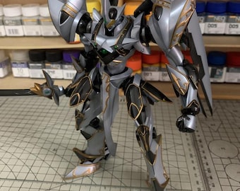 May include: A detailed model of a silver and black robot with gold accents, holding a sword. The robot has a complex design with multiple panels and joints. The model is on a cutting mat with the text "ArrowModelBuild".