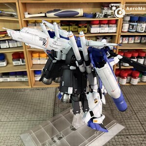 May include: A partially assembled model kit of a mecha robot, predominantly white, blue, and black. The model displays intricate details, including multiple thrusters and articulated components. It is displayed on a clear stand, with paint supplies visible in the background.