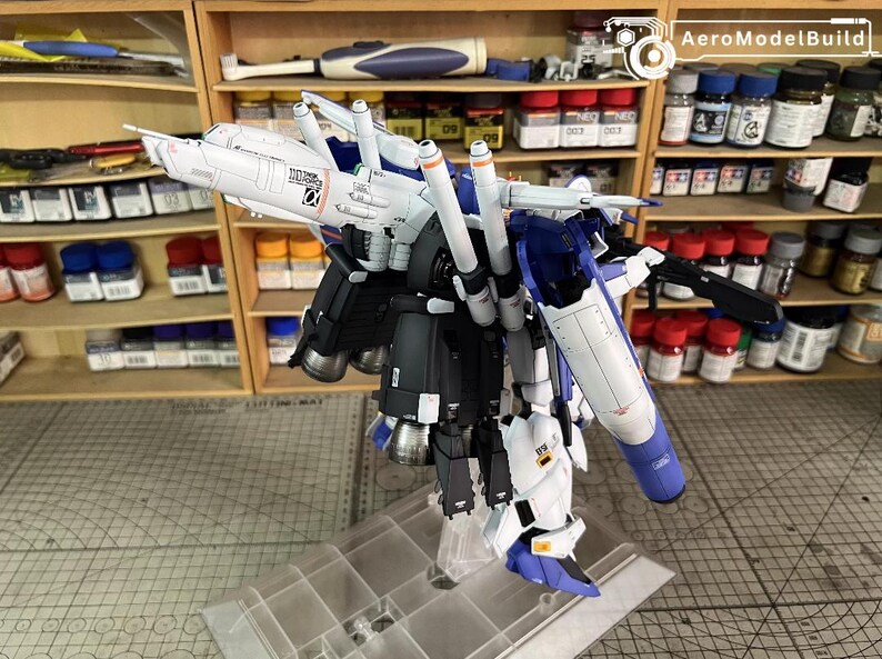 May include: A partially assembled model kit of a mecha robot, predominantly white, blue, and black. The model displays intricate details, including multiple thrusters and articulated components. It is displayed on a clear stand, with paint supplies visible in the background.