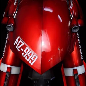 May include: A close-up of a red robot model with white accents and the text "NZ-999". The model has a glossy finish and features cylindrical components and metallic details. The AeroModeBuild logo is visible.