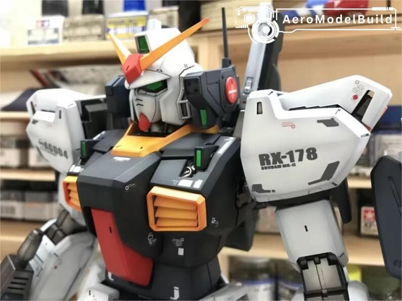 Op de afbeelding: Gedetailleerd model van een Gundam RX-178, met een wit, zwart, oranje en rood kleurenschema. Het model heeft ingewikkelde details, waaronder ventilatieopeningen, panelen en de tekst "RX-178" op de schouder. Het is een verzamelobject.