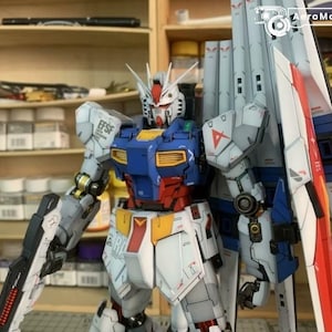 K&ouml;nnte beinhalten: Detailliertes Modell eines Gundam-Roboters mit einer Farbgebung aus Wei&szlig;, Blau, Rot und Gelb. Der Roboter hat gro&szlig;e Schulterpanzer und h&auml;lt eine Waffe. Das Modell steht vor Regalen mit verschiedenen Beh&auml;ltern.