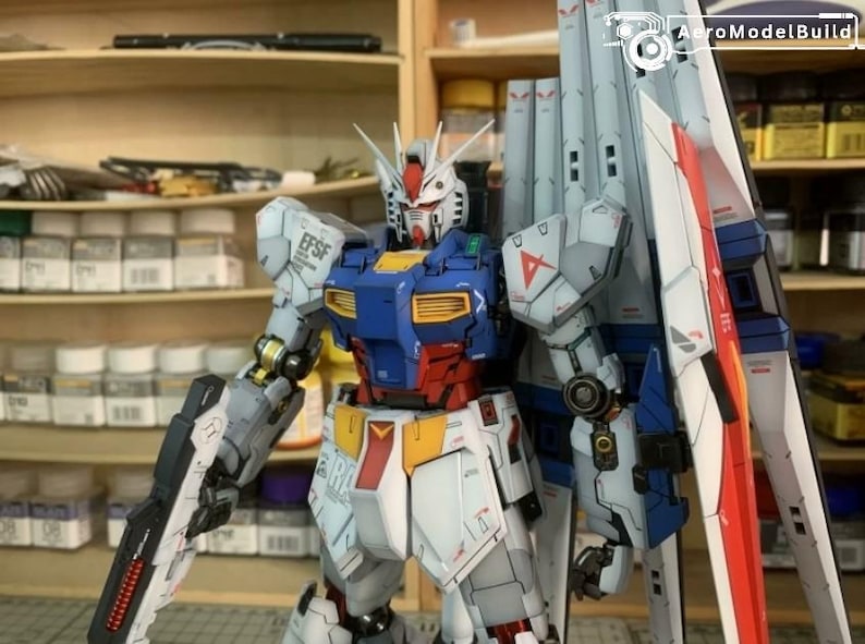 K&ouml;nnte beinhalten: Detailliertes Modell eines Gundam-Roboters mit einer Farbgebung aus Wei&szlig;, Blau, Rot und Gelb. Der Roboter hat gro&szlig;e Schulterpanzer und h&auml;lt eine Waffe. Das Modell steht vor Regalen mit verschiedenen Beh&auml;ltern.